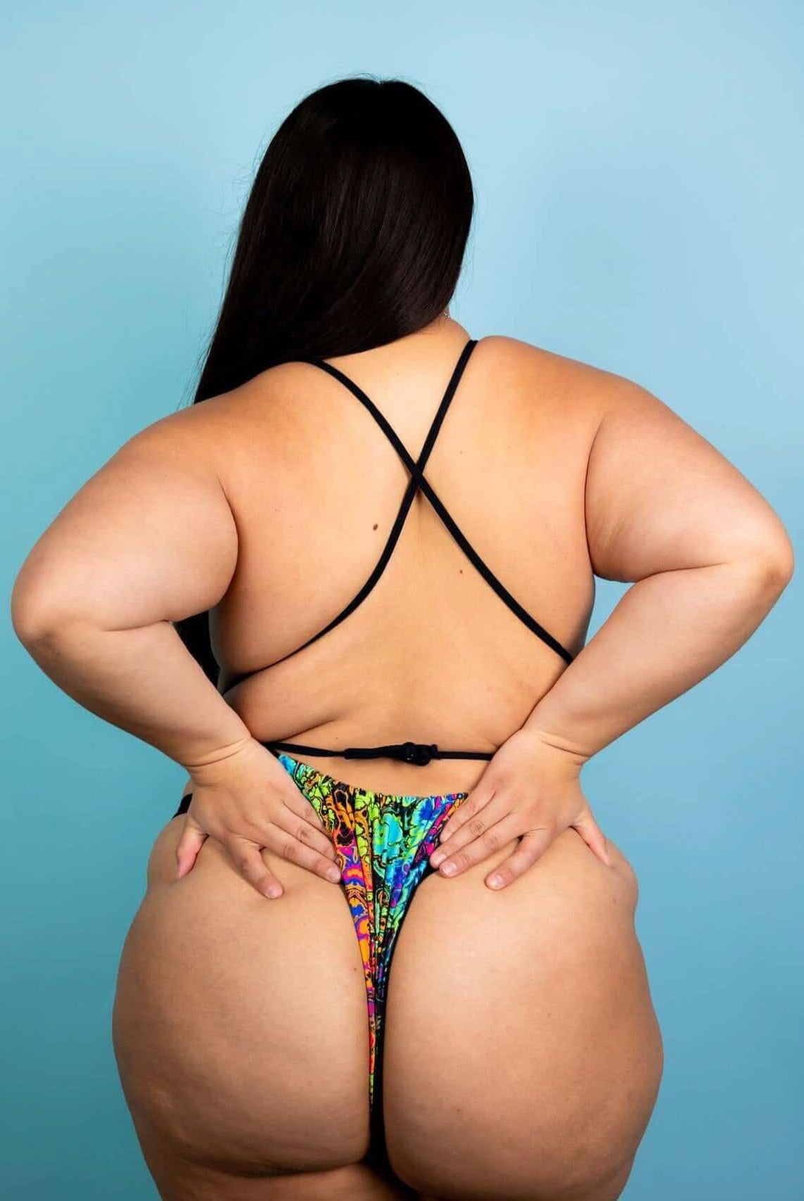 Back view of a model wearing the Lucidity Temptation Cheeky One Piece, showcasing its vibrant rainbow design and minimal coverage.