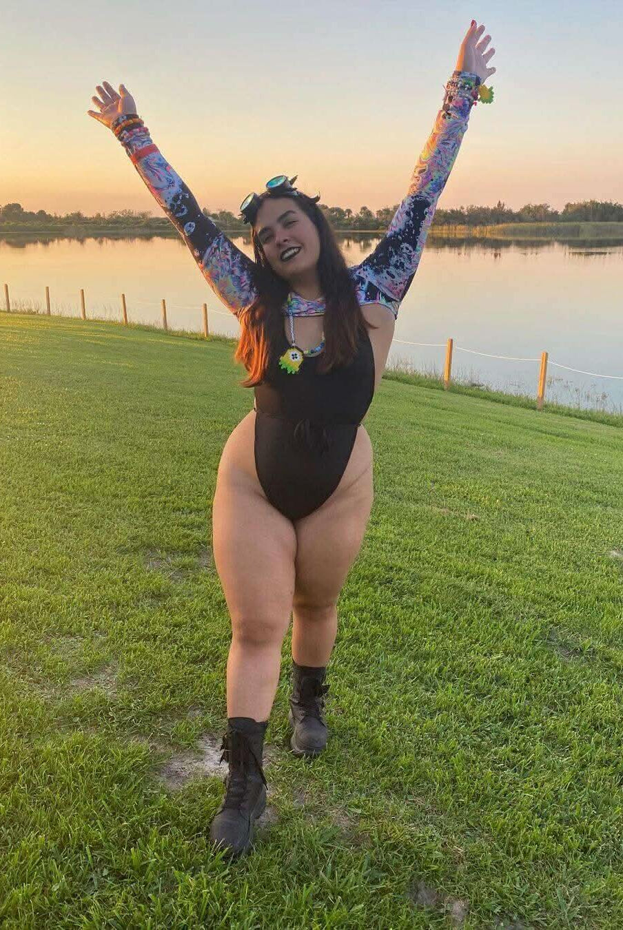 Woman in a black one-piece swimsuit with colorful sleeves, joyfully celebrating by the water at sunset.