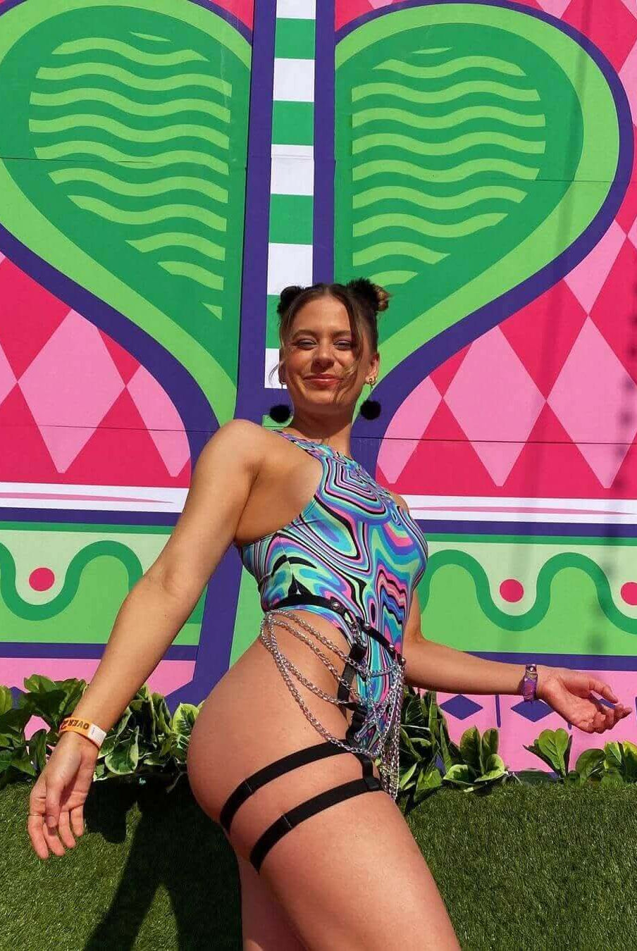 Woman wearing a colorful bodysuit with side chains, posing against a vibrant heart-patterned mural at a rave event.