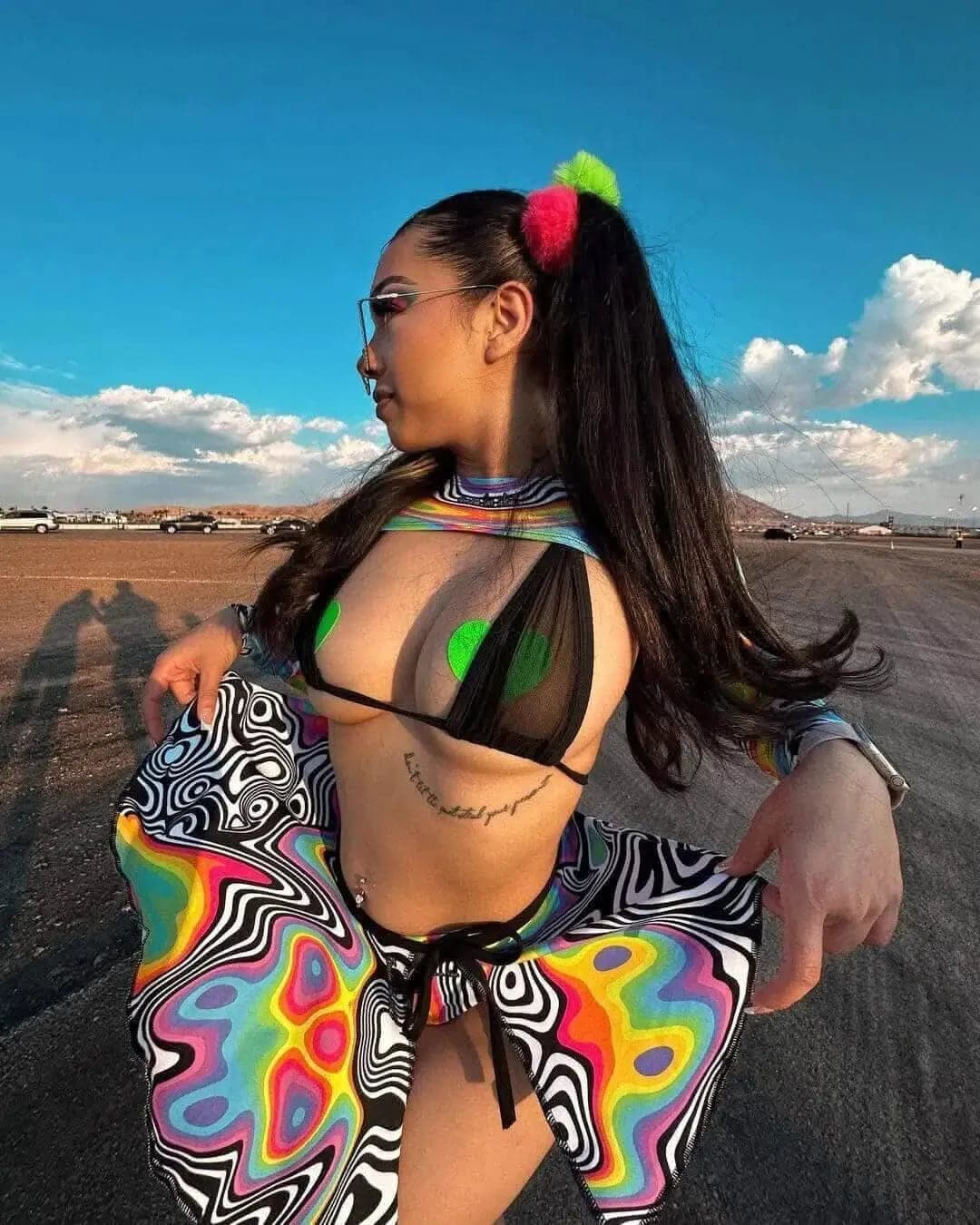 Woman wearing a black mesh bikini top and a vibrant psychedelic skirt at a festival, side view.