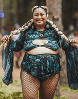 A smiling woman in the forest wears plus-size rave outfits, featuring a vibrant two-piece set with green and black patterns, accessorized with fishnet stockings and braided hair, embracing the festival vibe