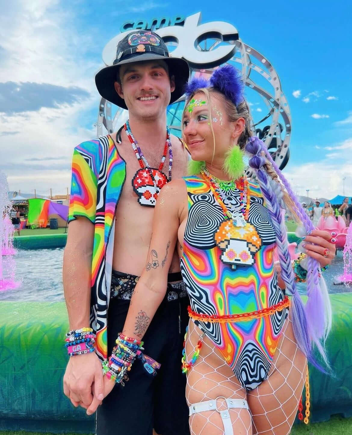 A couple poses at a festival, both wearing colorful psychedelic-patterned rave outfits. She wears a fishnet bodysuit with accessories, and he wears a matching shirt and hat