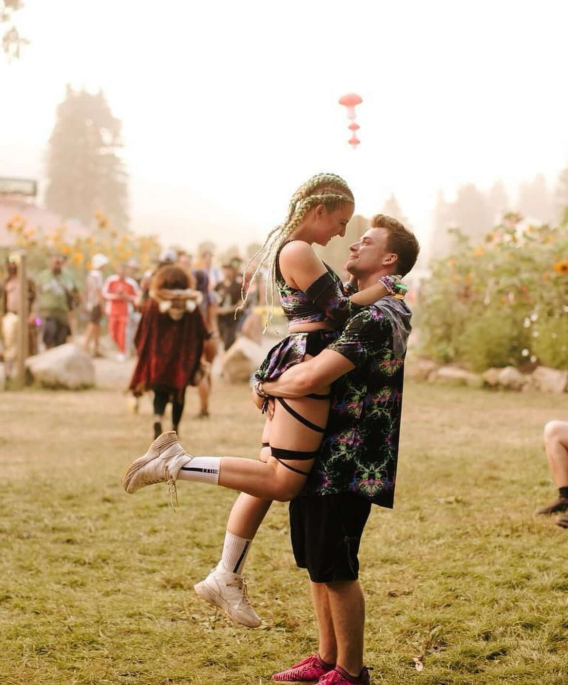 A couple embracing at a festival, with the woman being lifted off the ground by the man. The woman has braided hair and is wearing festival attire, while the man is dressed in a colorful shirt and shorts
