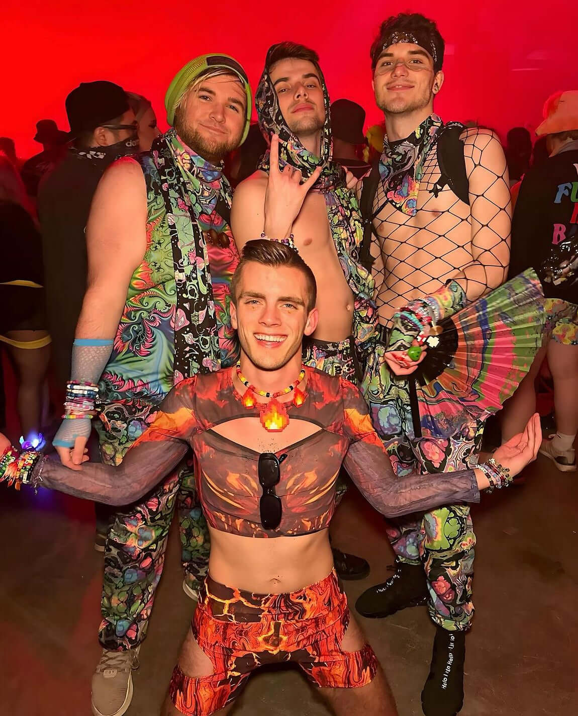 A group of festival-goers in vibrant men's rave clothes, featuring colorful patterns and fishnet tops, poses confidently in a lively club setting