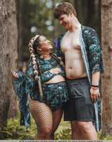 A joyful couple dressed in matching festival outfits, the woman in a plus-size green and black patterned two-piece set with fishnet stockings, and the man in matching shorts and an open robe, sharing a happy moment in the forest.