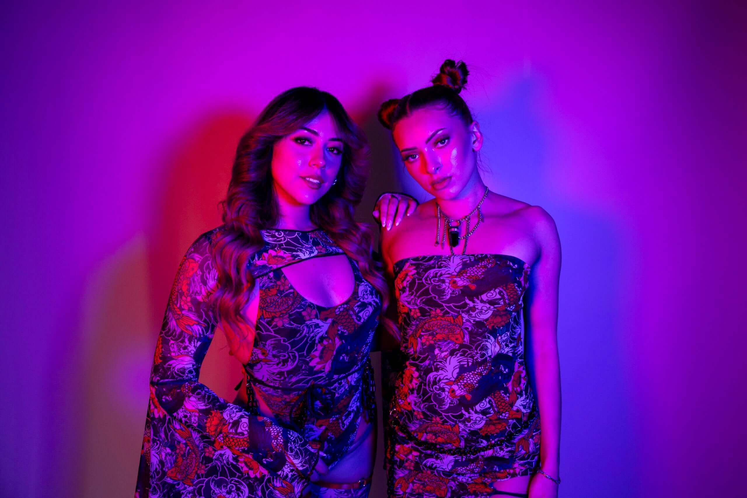 Two women in colorful rave outfits posing under vibrant purple lighting.