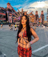 Stylish woman in a red and black rave outfit with intricate patterns and chain accents, confidently posing at a vibrant festival scene