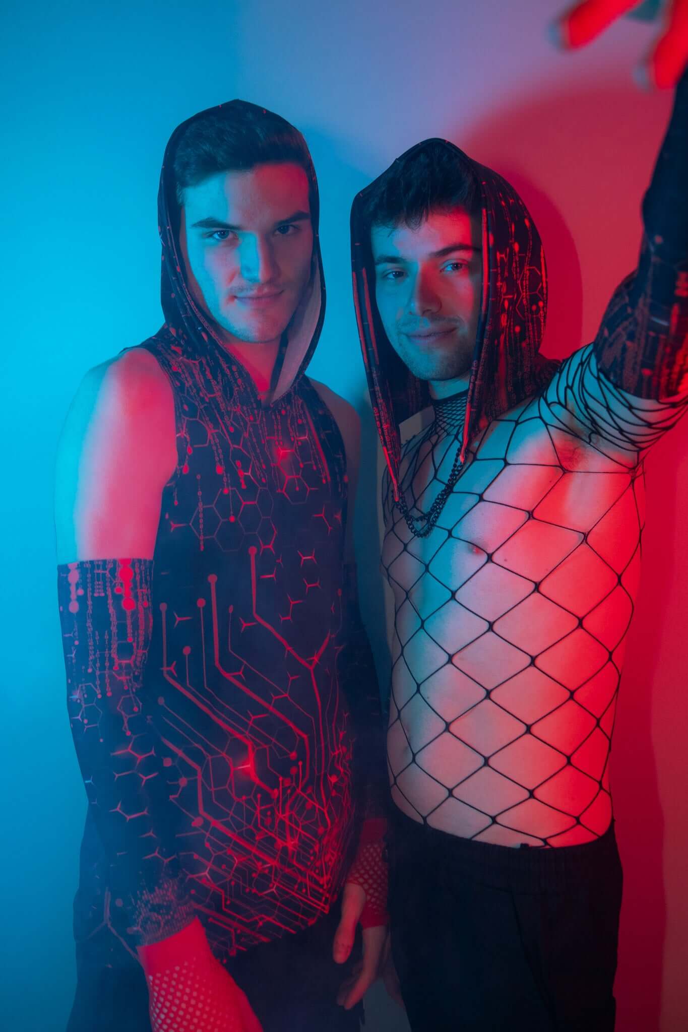 Two men wearing stylish rave gear, including a hooded sleeveless top with a futuristic red and black circuit design and a fishnet top, standing in dramatic blue and red lighting.