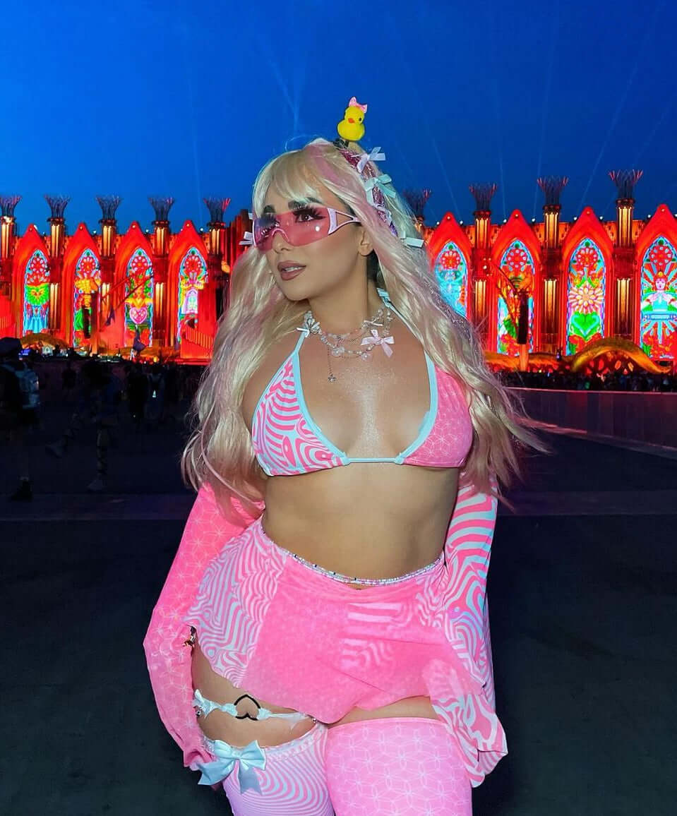 A festival-goer in a vibrant women's rave outfit poses confidently. She wears a pink patterned bikini set with matching accessories, thigh-high socks, and sunglasses, set against a colorful festival backdrop at night