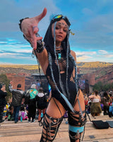 A festival-goer in a black rave bodysuit poses confidently with braided hair and accessories, set against a scenic outdoor venue