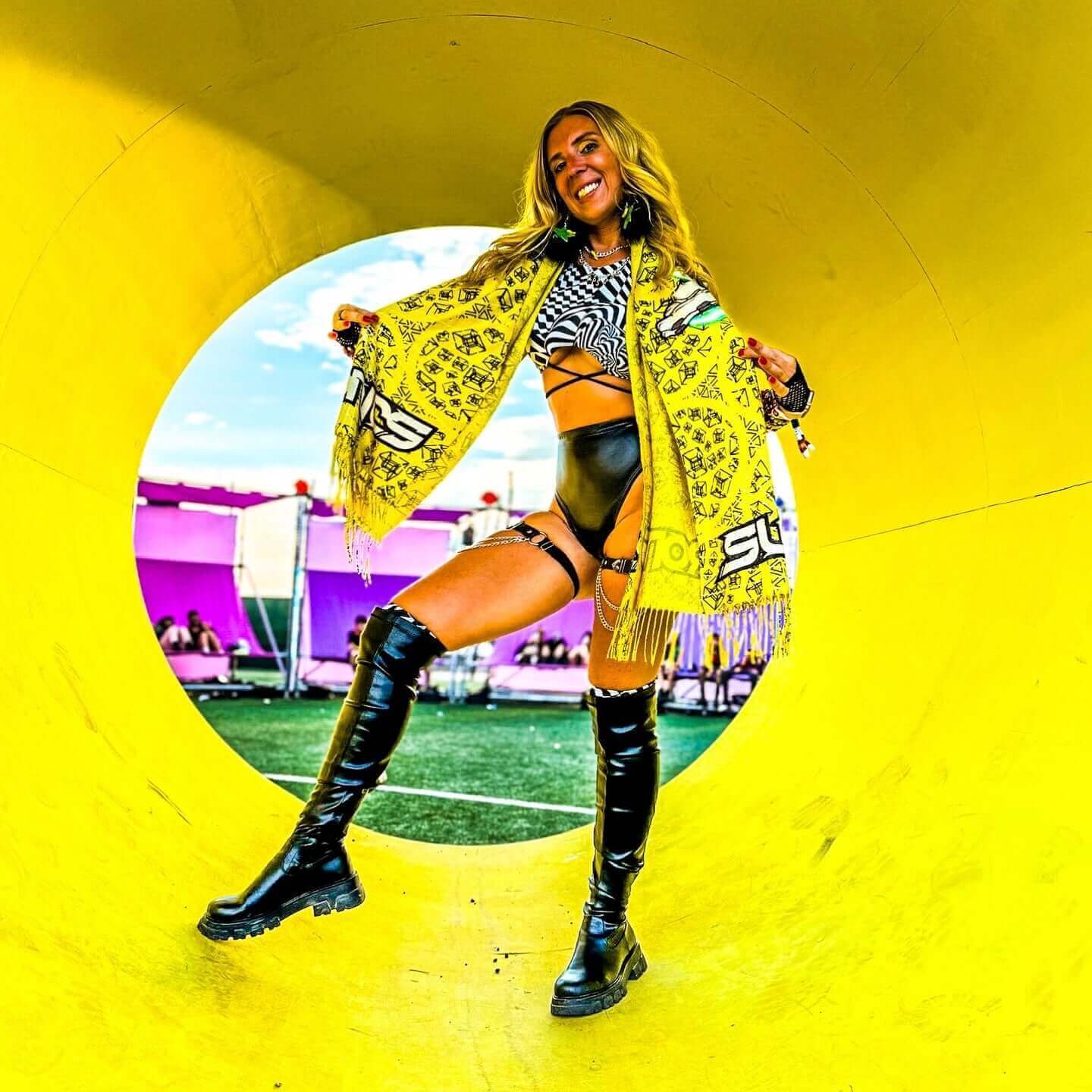 Woman in stylish rave outfit poses with a vibrant scarf in a colorful festival setting.