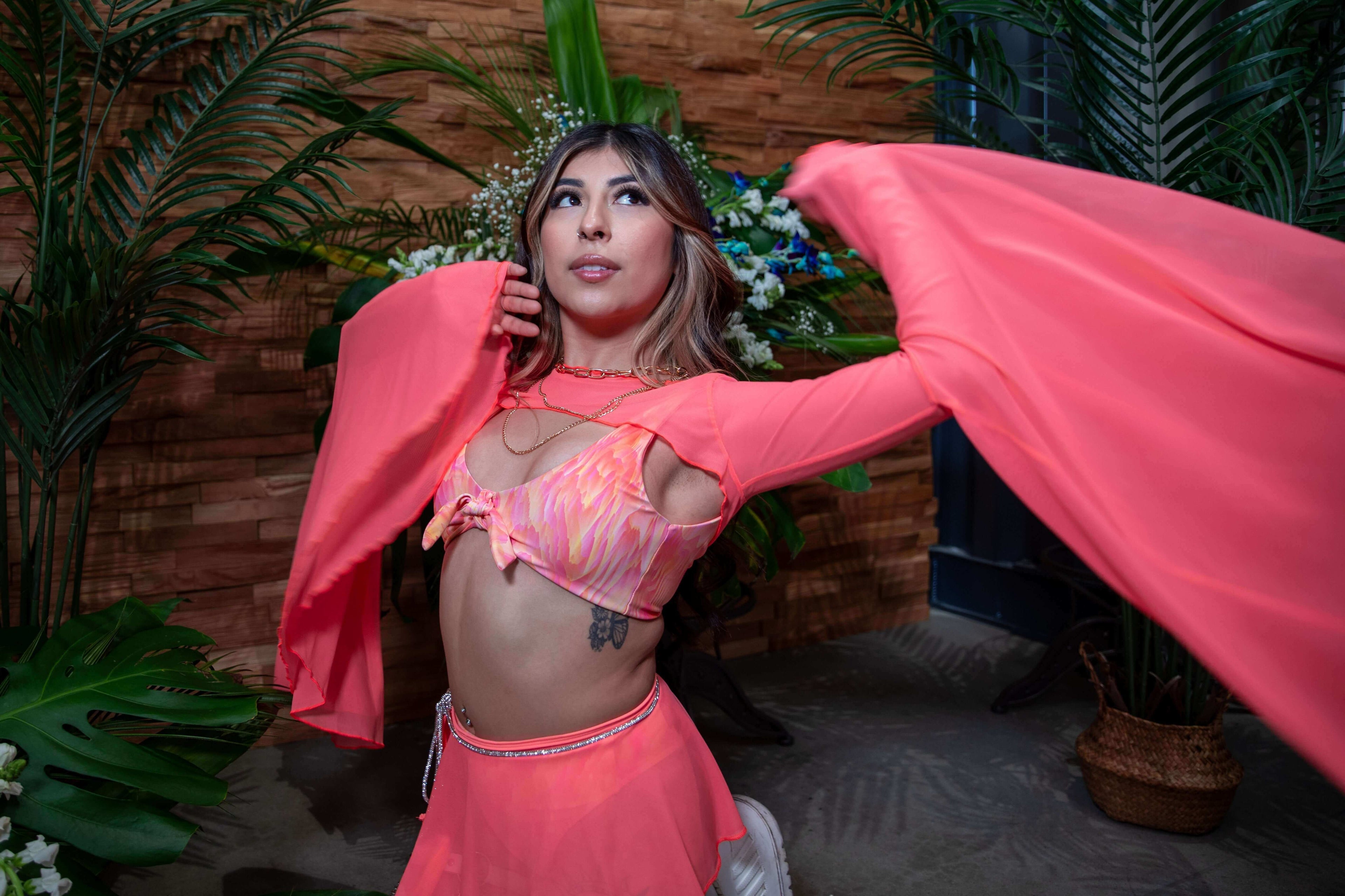 Woman in vibrant coral outfit with flowing sleeves, posing in a lush tropical setting for festival aesthetics.