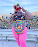 Confident woman in a colorful rave outfit enjoying a festival. She sports a vibrant bodysuit with psychedelic patterns, pink leggings, and fluffy rainbow accessories, posing playfully against a backdrop of festival grounds and mountains