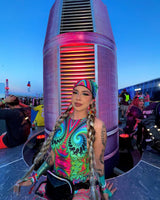 rave girl in a bright colorful hypnotic rave outfit with matching accessories sits in from of art structure at a music festivals