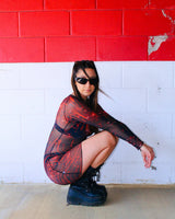 rave girl in a red and black mesh rave dress kneels in front of a red and white wall with sunglasses