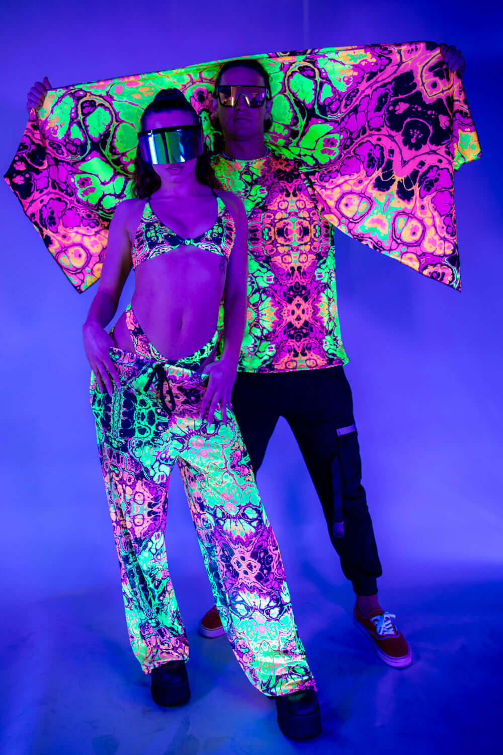 Model wearing a vibrant rave bodysuit with colorful patterns and black mesh sleeves, holding out sheer black fabric attached to the outfit