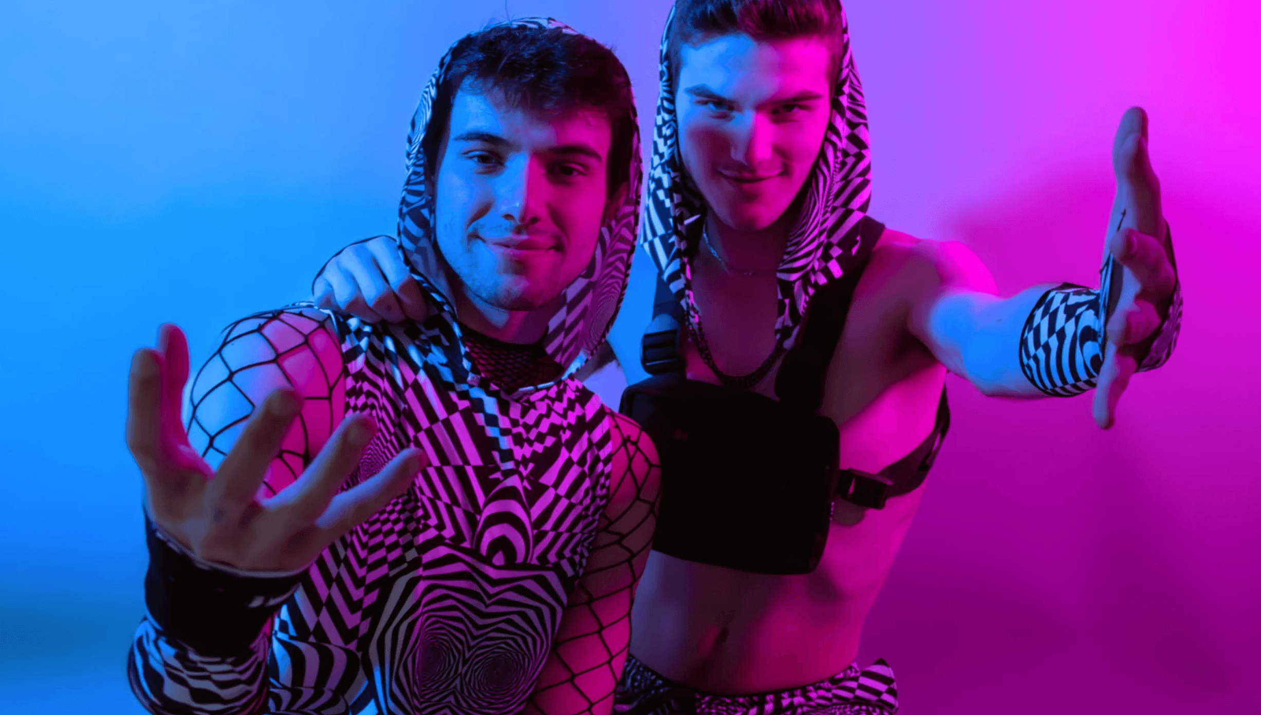 Two men in matching black and white geometric rave outfits pose under vibrant blue and pink lighting, showcasing trendy festival fashion