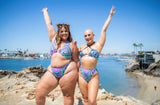 Two women in colorful psychedelic rave outfits pose joyfully by the beach, showcasing festival fashion and body positivity.