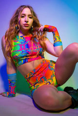 Model in a colorful, psychedelic-patterned rave outfit with matching gloves, posing confidently with vibrant lighting