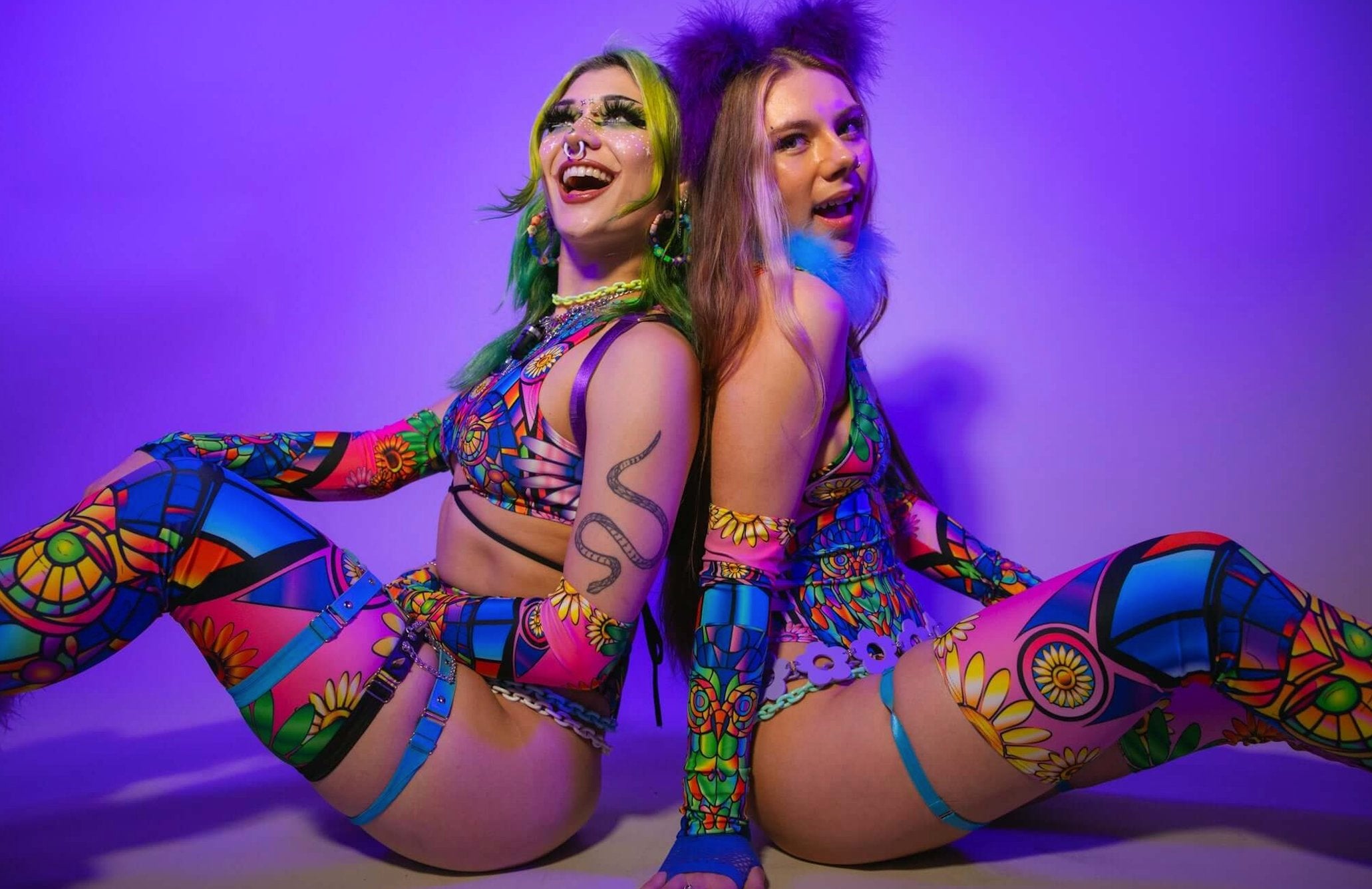 Two models in vibrant, colorful rave outfits featuring intricate floral and geometric patterns, sitting back-to-back with a purple background