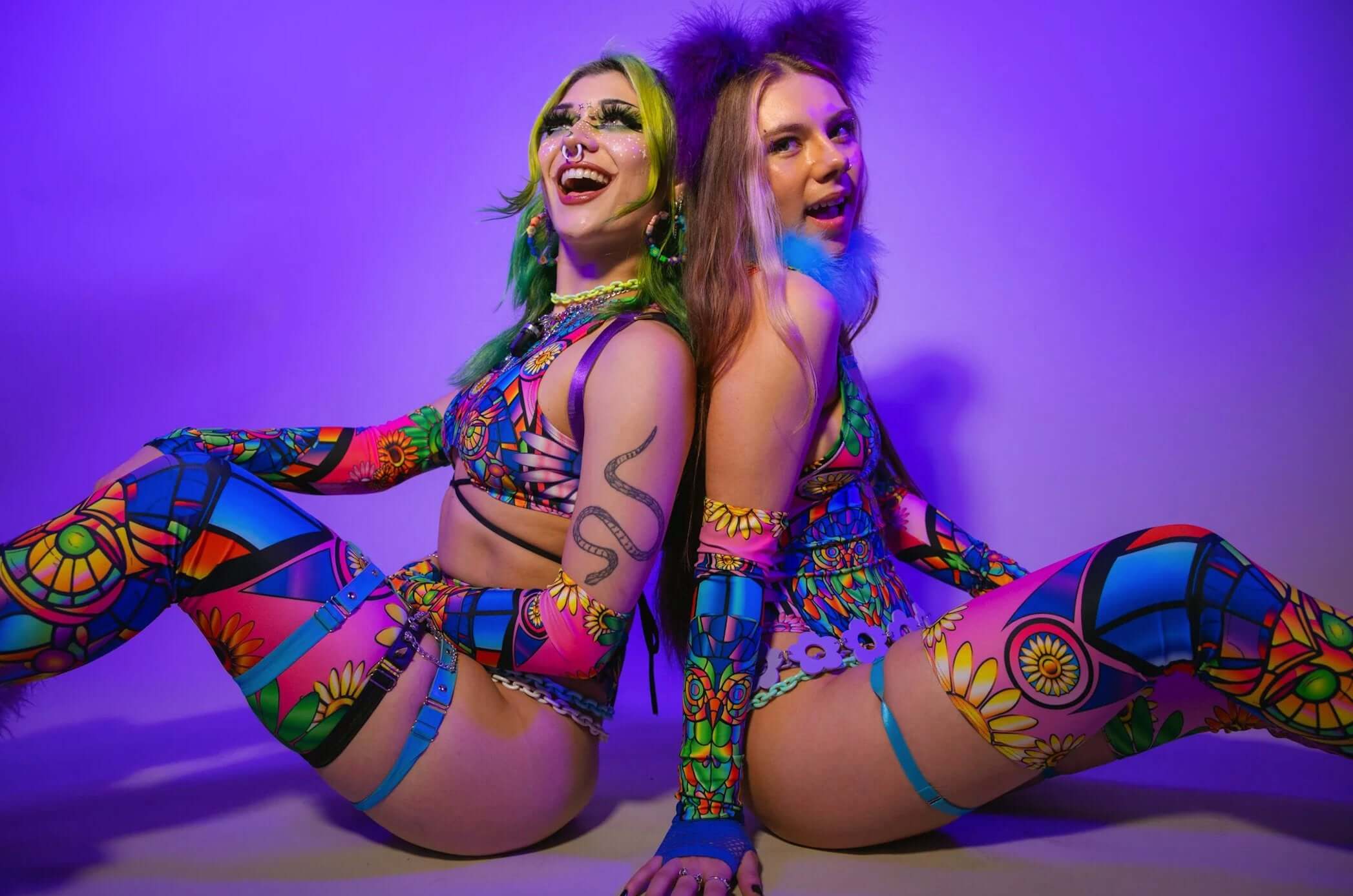 Two women in vibrant, psychedelic rave outfits with colorful patterns and playful accessories sit back-to-back, embodying festival fashion