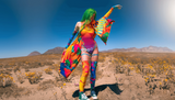 Green-haired woman in a colorful, psychedelic rave outfit poses in a desert landscape, showcasing vibrant festival fashion