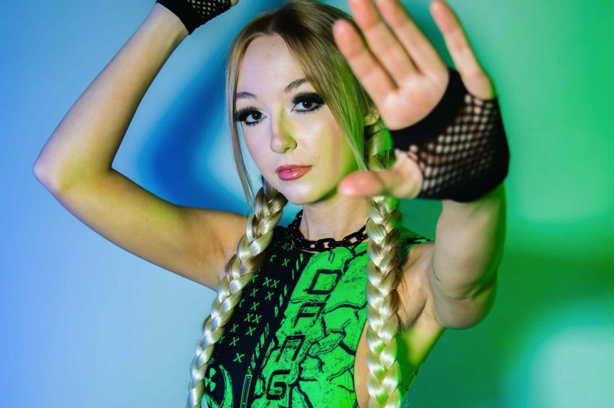 A woman in a vibrant green and black rave outfit with long blonde braids, posing confidently with hands raised, in an edgy, colorful setting