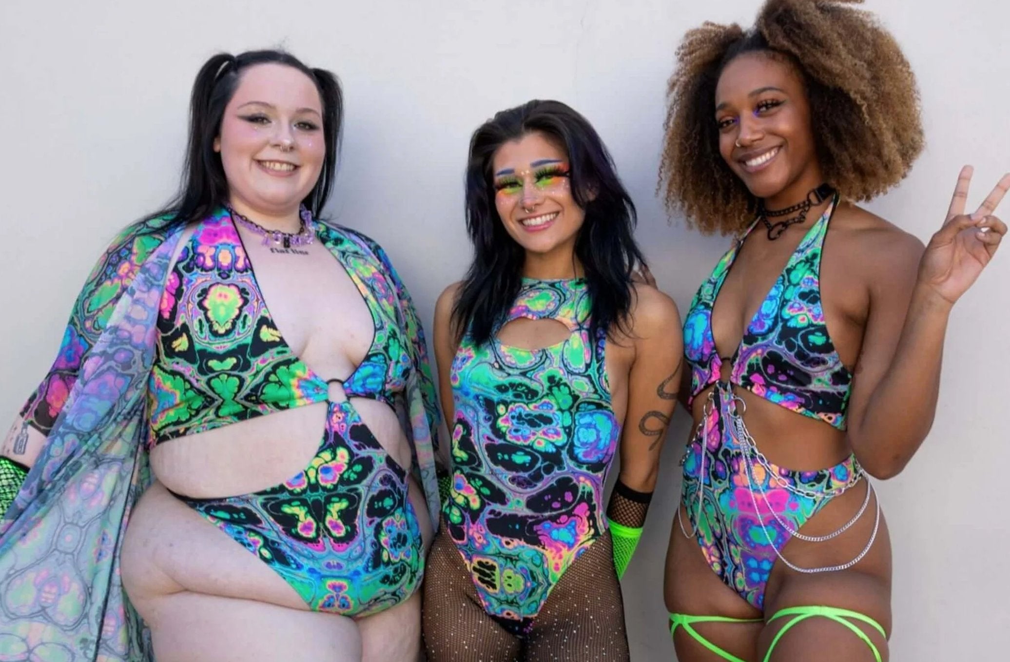 Three women in vibrant, multicolored cut-out rave bodysuits, smiling and posing together, showcasing diverse body types and styles