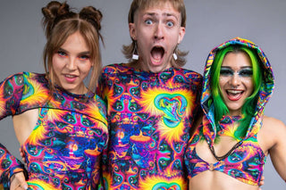 Three people in vibrant, psychedelic-patterned rave outfits pose together; one making a surprised face, flanked by two smiling women, one with a hood and one with buns in her hair.