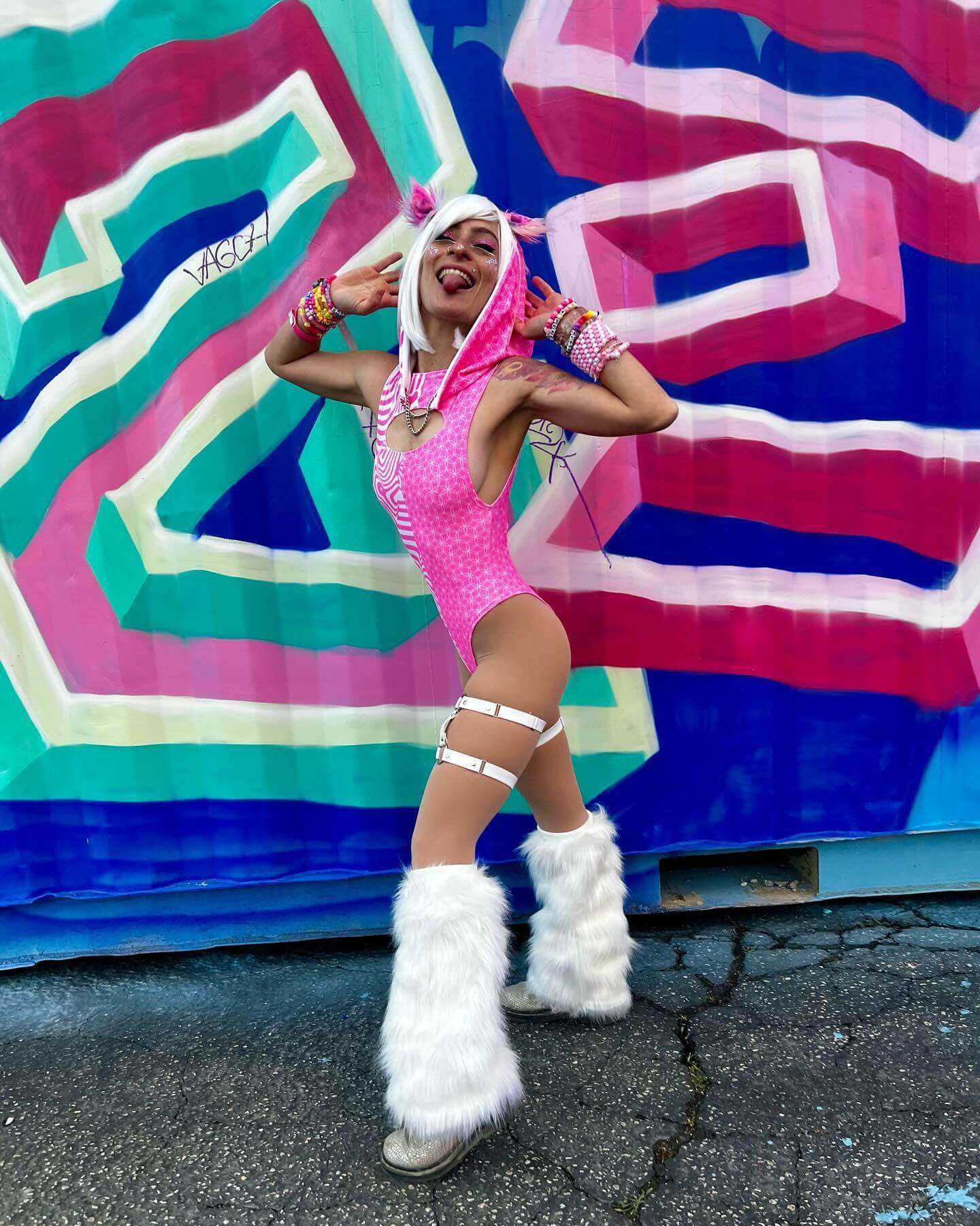 A woman playfully poses in front of a mural, wearing a pink and white bodysuit with a matching hood, and fluffy white boots.