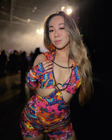 Rave enthusiast in a vibrant, multi-colored outfit poses with allure at a dimly lit music event, embodying the rave spirit