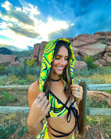 rave girl wears bright yellow and green hooded festival outfit with neon makeup against the backdrop of red rocks and blue skies