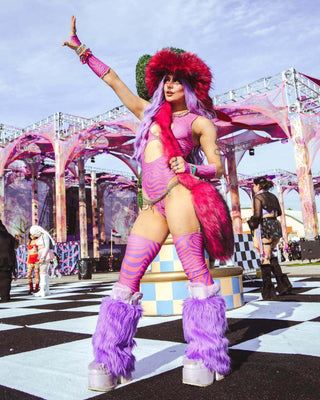 A photo of a woman wearing a pink and purple swirled bodysuit with two keyhole cutouts. She is wearing matching leg sleeves and gloves with a furry hat. She is at a music festival.