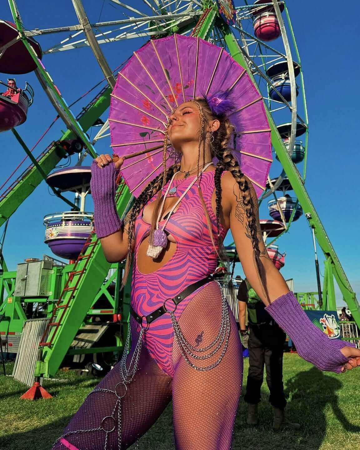 Young woman in a vibrant rave outfit with a purple parasol, posing near a colorful Ferris wheel at an outdoor festival.