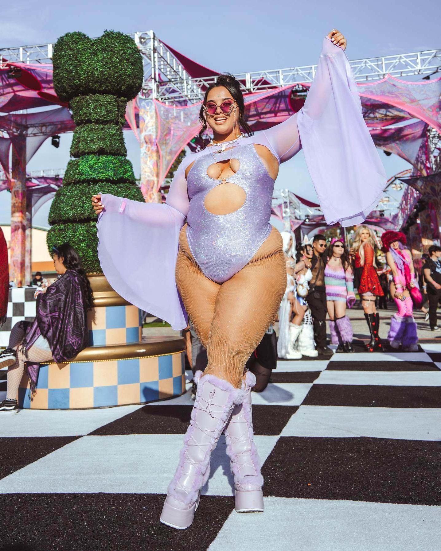 Model in lavender holo keyhole bodysuit with flowing sleeves, posing at a lively outdoor rave event.