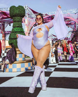 Confident rave girl in a sparkling lilac bodysuit with matching boots dances at a lively festival atmosphere.