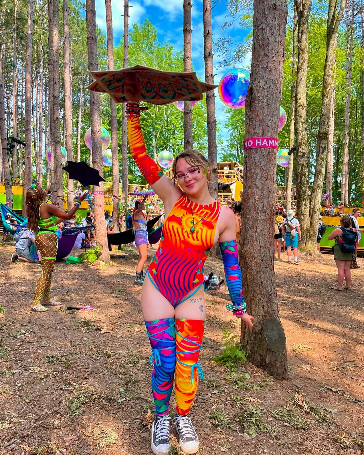 A woman in a colorful rave outfit poses in the woods, holding a decorated piece above her head, surrounded by festival-goers