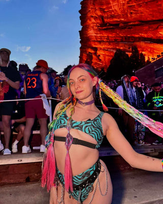 A woman wearing a blue and black swirled crop top with a matching bikini bottom. She poses at a music festival.