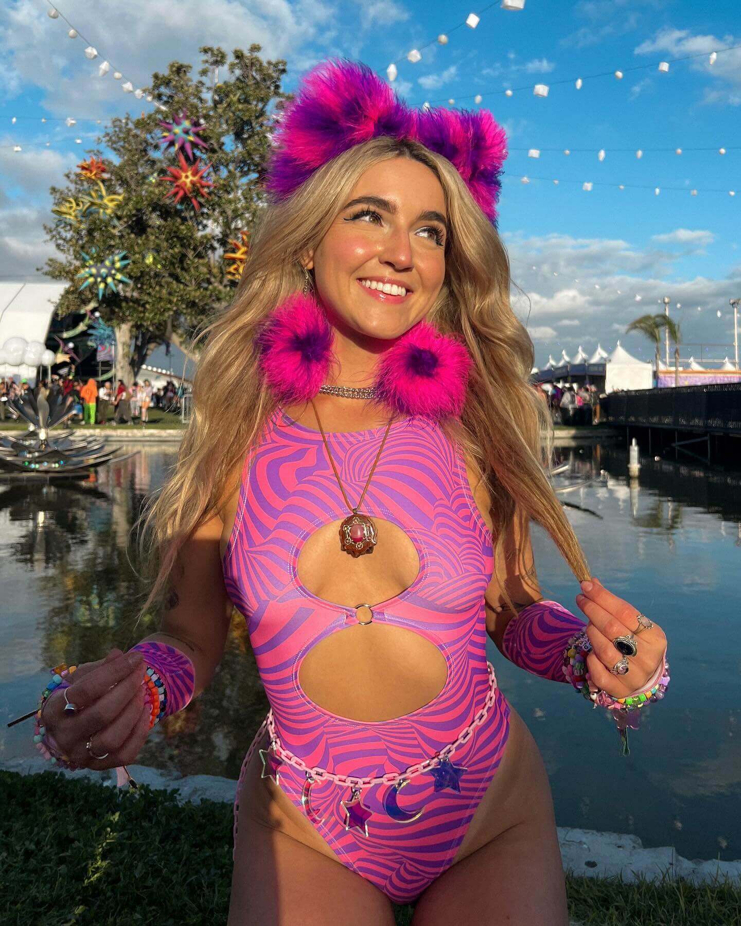 A photo of a woman wearing a pink and purple swirled bodysuit with two keyhole cutouts. She is wearing fluffy pink and purple accessories. She is standing in front of the water and smiling. 