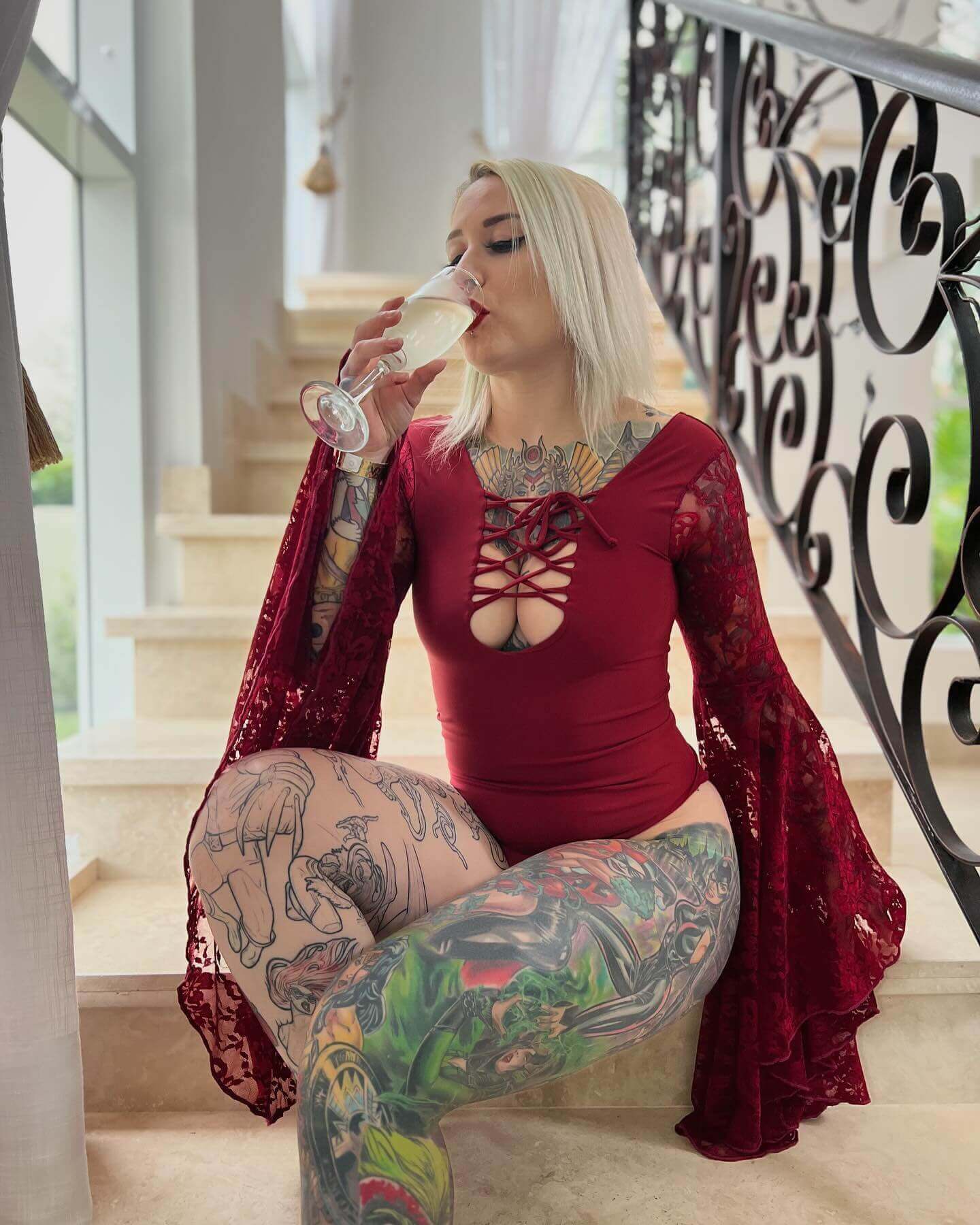 Elegant scene with a tattooed woman in a red lace-sleeved rave bodysuit from Freedom Rave Wear, sipping champagne