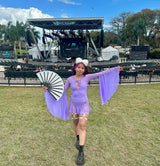 Woman wearing a lavender Freedom Rave Wear bodysuit with mesh sleeves, garters, and a fan, posing in front of an outdoor concert stage on a sunny day