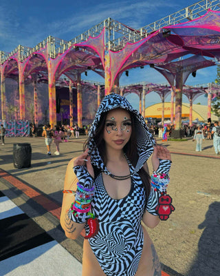 A woman at a festival wearing a black and white striped hooded outfit, adorned with colorful bracelets and face paint, stands before a vibrant stage structure.