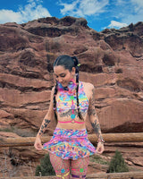 A woman standing in front of rocky terrain wearing a rainbow crop top and matching skirt.