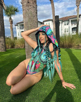 rave girl poses in front of lush green backdrop wearing colorful trippy print rave outfit and matching hood