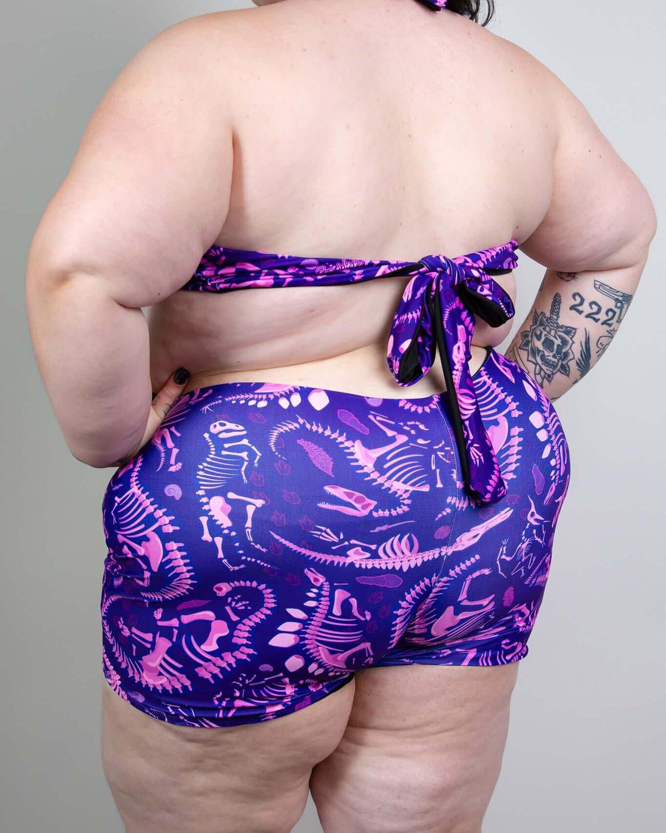 Back view of a plus-size model in a purple dino fossil romper with a halter tie, perfect for rave outfits.