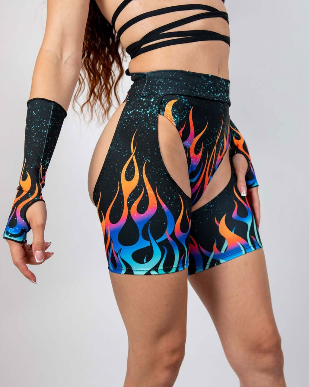 Model wearing Blaze Chaps in fiery flame graphics, showcasing high-waisted fit and open hip cutouts, ideal rave outfits.