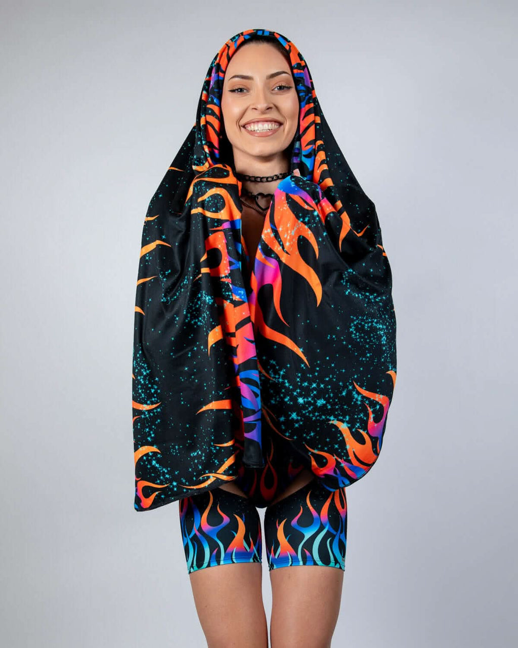 Smiling model wearing Blaze Festival Scarf styled as a hood, showcasing neon flames and cosmic print. Perfect for rave outfits.