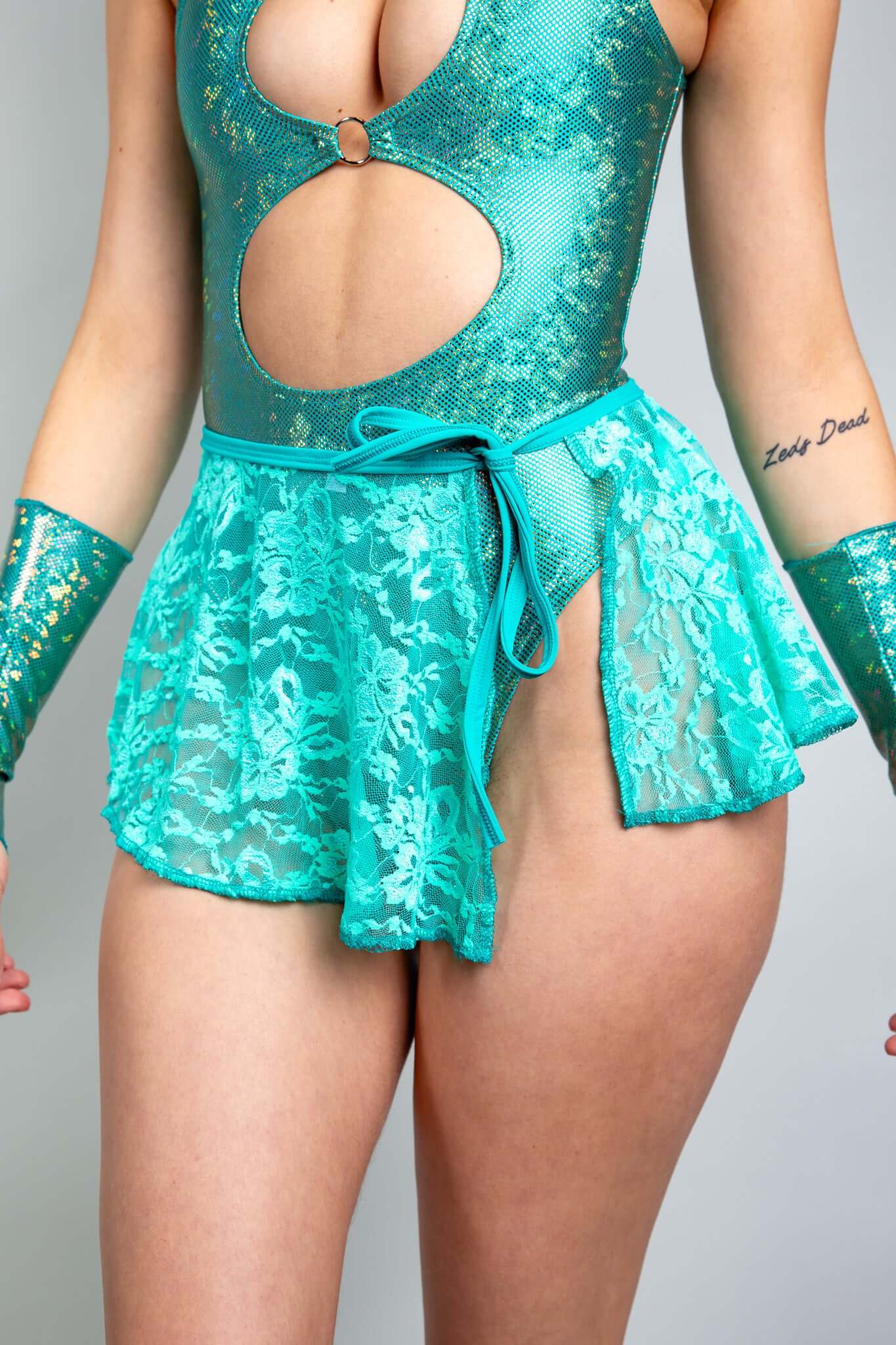 An up close photo of a woman wearing a teal, holographic bodysuit with two keyhole cutouts and a teal lace skirt that ties on the side.