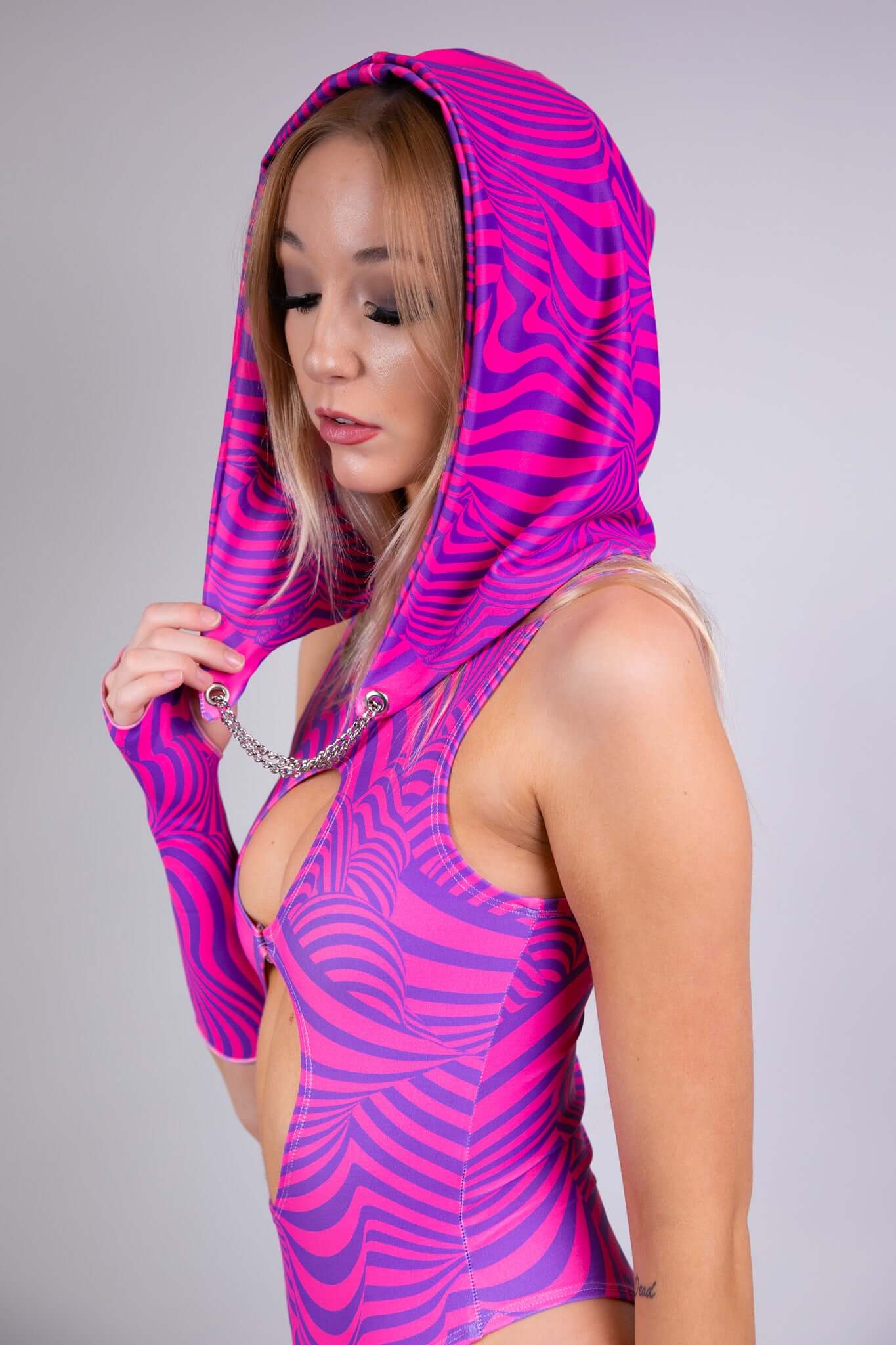 A woman with a hood and sleeveless bodysuit in pink and purple swirl patterns, featuring cut-out details. She has blonde hair and makeup.