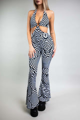 A woman wearing a black and white geometric printed jumpsuit with bell bottoms. The jumpsuit is a halter neck with cutouts on either side and an o-ring detail.